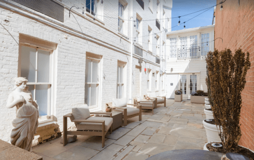 A serene courtyard with stone paving, white walls, outdoor seating, and a statue, surrounded by potted plants.
