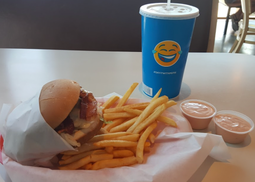 A burger with bacon and lettuce, served with fries and two dipping sauces, alongside a blue drink cup.