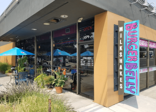 Exterior of Burger Belly restaurant with outdoor seating, umbrellas, and a colorful sign for milkshakes and burgers.