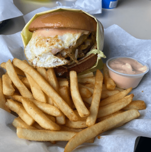 A burger with an egg and toppings, served with a side of crispy French fries and a small cup of sauce.