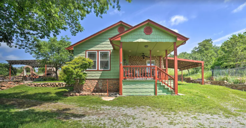 A charming green house with a red porch, surrounded by trees and a grassy yard under a blue sky.