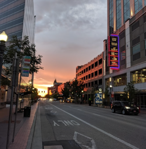 A city street at sunset, with buildings on either side and a colorful sky reflecting on the horizon.