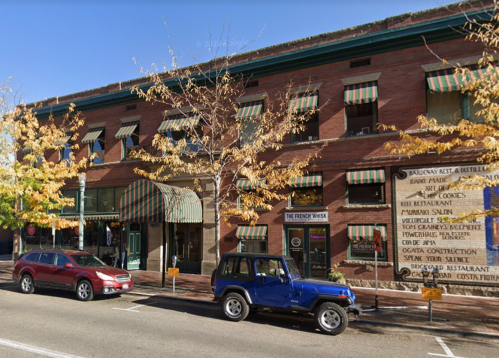 A brick building with colorful awnings, featuring shops and restaurants, alongside parked cars on a tree-lined street.