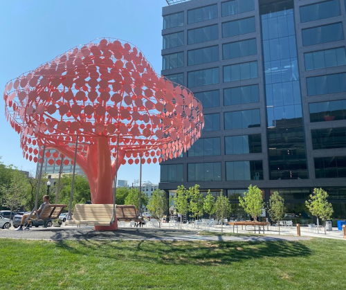 A large pink tree sculpture stands in a park with benches, surrounded by greenery and a modern building in the background.