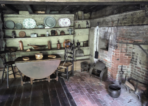 A rustic kitchen with wooden shelves, pots, and a brick fireplace, featuring a round table and various kitchen tools.