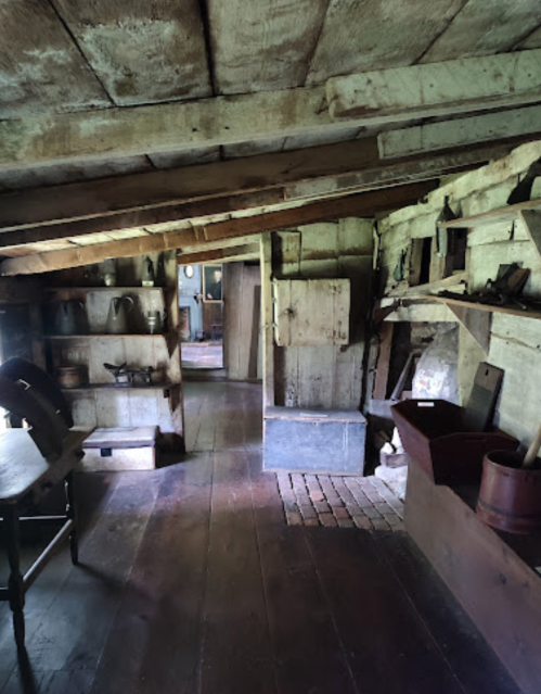 A rustic interior of an old wooden house, featuring shelves, a table, and various vintage kitchen items.