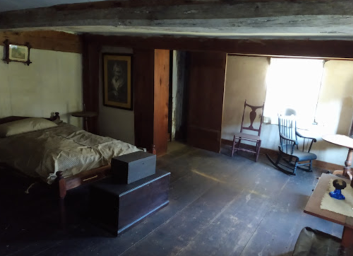 A rustic bedroom with a bed, rocking chair, table, and window, featuring wooden beams and simple decor.