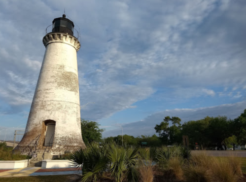 A tall, white lighthouse stands against a cloudy sky, surrounded by greenery and a pathway.