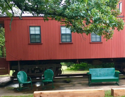 A red train car on wheels, with green chairs and benches nearby, surrounded by trees and grass.