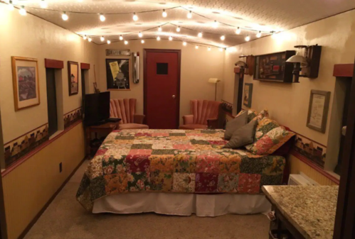 Cozy room with a bed, patterned quilt, two chairs, TV, and string lights on the ceiling. Warm, inviting atmosphere.