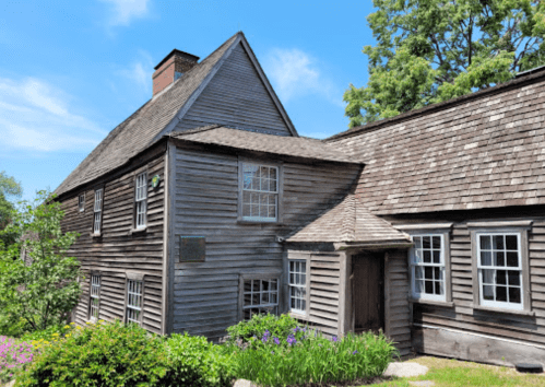 Historic wooden house with a steep roof, surrounded by greenery and colorful flowers under a clear blue sky.