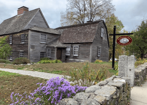 Historic wooden house with a stone pathway and purple flowers in the foreground, featuring a sign nearby.