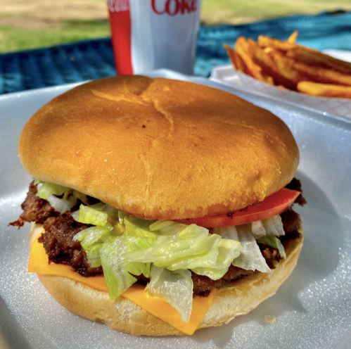 A close-up of a cheeseburger with lettuce, tomato, and a soft bun, served with fries and a soda in the background.