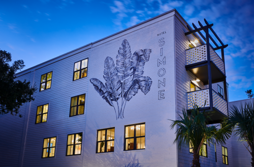 A hotel building at dusk, featuring a large mural of tropical leaves and illuminated windows.