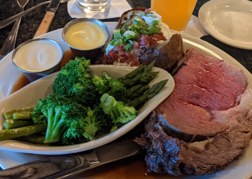 A plate featuring prime rib, baked potato with toppings, and a side of broccoli and asparagus.