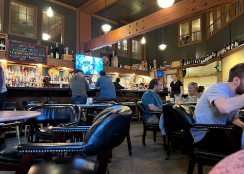 A cozy bar interior with patrons at the counter and tables, featuring wooden beams and a TV displaying sports.