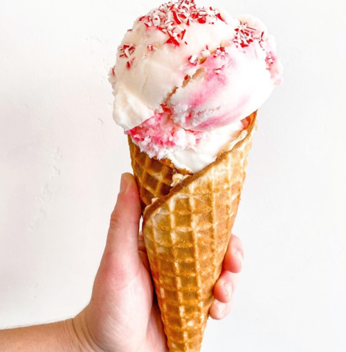 A hand holding a waffle cone filled with pink and white ice cream topped with red sprinkles against a white background.