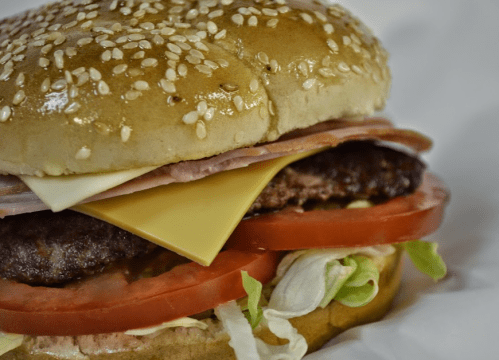 A close-up of a sesame seed bun burger with lettuce, tomato, cheese, ham, and a beef patty.