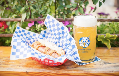 A hot dog in a basket with blue checkered paper, alongside a glass of beer on a wooden table with flowers in the background.