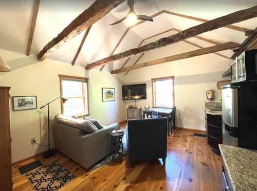 Cozy living space with wooden beams, a sofa, TV, dining area, and kitchen appliances in a bright, inviting room.