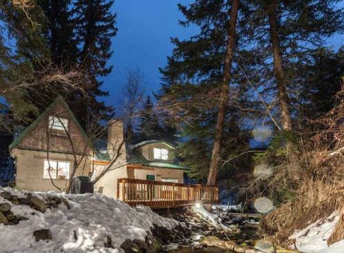 A cozy cabin by a snowy creek, surrounded by tall trees, illuminated at dusk.