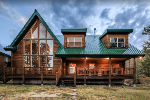 A large wooden cabin with a green metal roof, featuring a spacious porch and large windows, set against a cloudy sky.