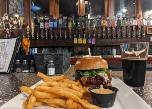 A burger with lettuce and sauce, fries, and a glass of dark beer on a table in front of a bar with many taps.