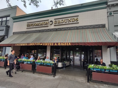 A café with outdoor seating, "Uncommon Grounds," featuring a striped awning and patrons enjoying coffee and bagels.