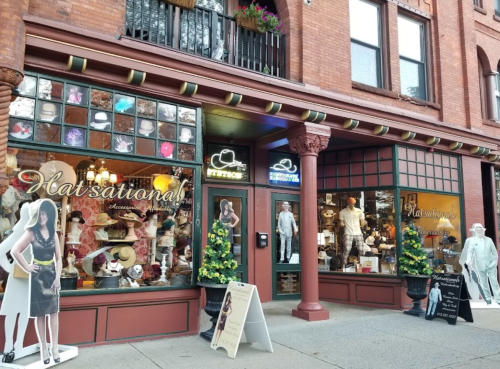 A storefront named "Hatsational!" featuring hats, mannequins, and decorative plants in a historic building.