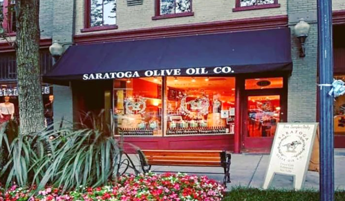 Exterior of Saratoga Olive Oil Co. store, featuring large windows and a sign, surrounded by flowers and greenery.