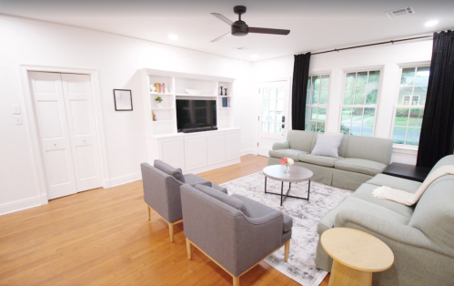 A modern living room with a light color scheme, featuring a TV unit, sofas, a coffee table, and large windows.
