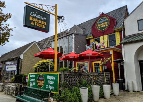 A colorful restaurant exterior with umbrellas, signage for burgers and fries, and a bench in front.