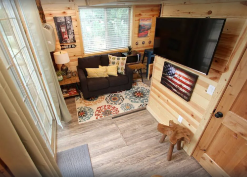 Cozy interior of a wooden cabin with a sofa, TV, colorful rug, and rustic decor, featuring large windows and natural light.
