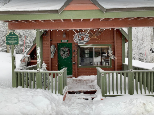 A cozy wooden cabin covered in snow, with green accents and festive wreaths on the door.