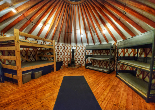 Interior of a yurt featuring wooden bunk beds, metal shelves, and a wooden floor with a circular ceiling design.