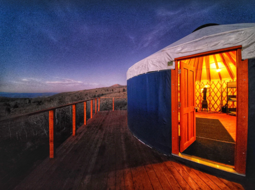 A cozy yurt with an open door, set on a wooden deck against a twilight sky and rolling hills.