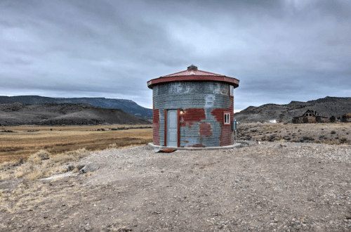 A small, round, corrugated metal building with a red and gray exterior, set against a cloudy sky and mountainous landscape.