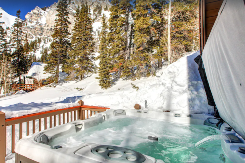 A hot tub on a snowy deck surrounded by tall pine trees and mountains in a winter landscape.