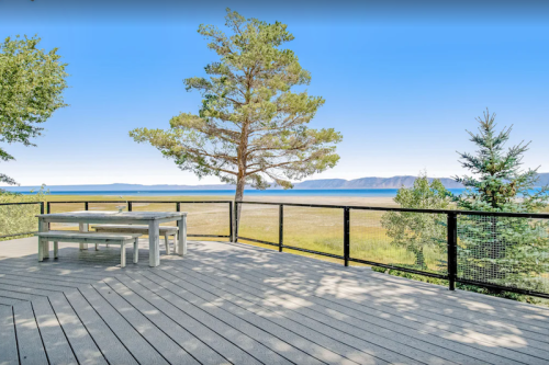 A wooden deck with a table overlooks a serene lake and distant mountains, surrounded by trees.