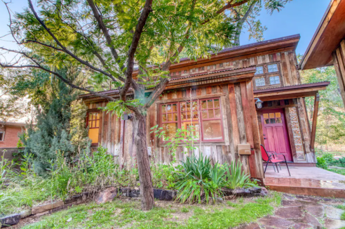 A rustic wooden house surrounded by greenery and trees, featuring large windows and a welcoming entrance.