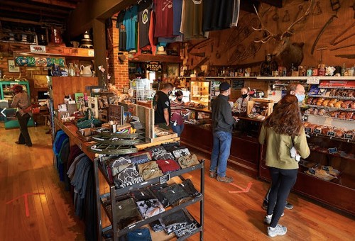 A cozy shop interior with clothing, snacks, and customers browsing at the counter. Wood accents and rustic decor are visible.