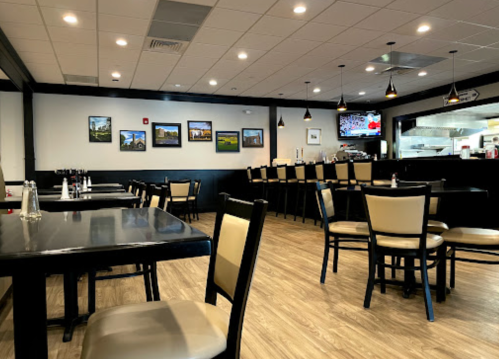 Interior of a restaurant featuring black and beige seating, tables, and wall art, with a visible kitchen area.