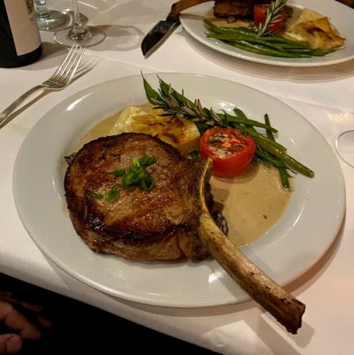 A plated meal featuring a ribeye steak, roasted tomato, green beans, and creamy potatoes, garnished with herbs.