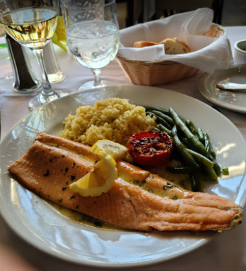 A plate of salmon with lemon, couscous, green beans, and a roasted tomato, accompanied by glasses of wine and water.