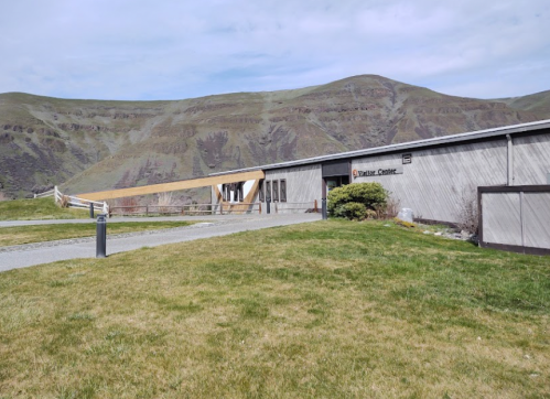 A modern building with a wooden ramp, set against a backdrop of green hills under a cloudy sky.