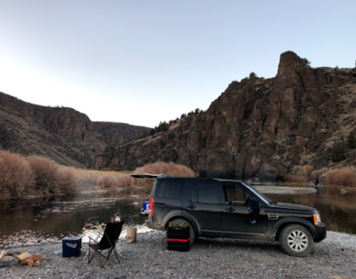 A black SUV parked by a river, with a camping chair and supplies nearby, surrounded by rocky cliffs and sparse vegetation.