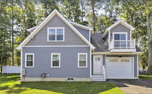 A modern gray house with a garage, white trim, and a balcony, surrounded by trees and a green lawn.