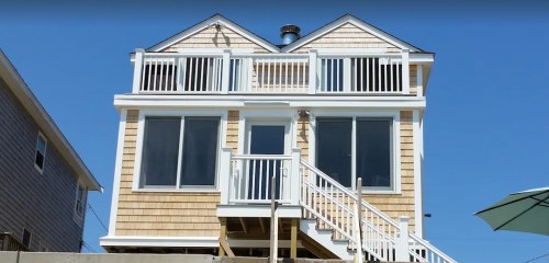 A two-story beach house with wooden siding, large windows, and a staircase leading to the entrance, under a clear blue sky.