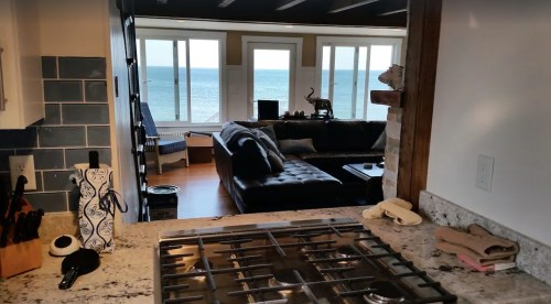 View from a kitchen with a gas stove, looking into a living room and out to a scenic ocean view through large windows.