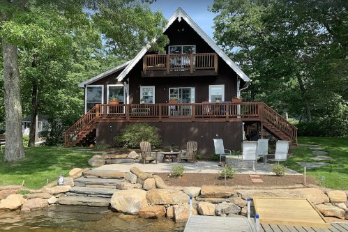 A cozy brown cabin with balconies, surrounded by greenery and a stone patio by a lake.